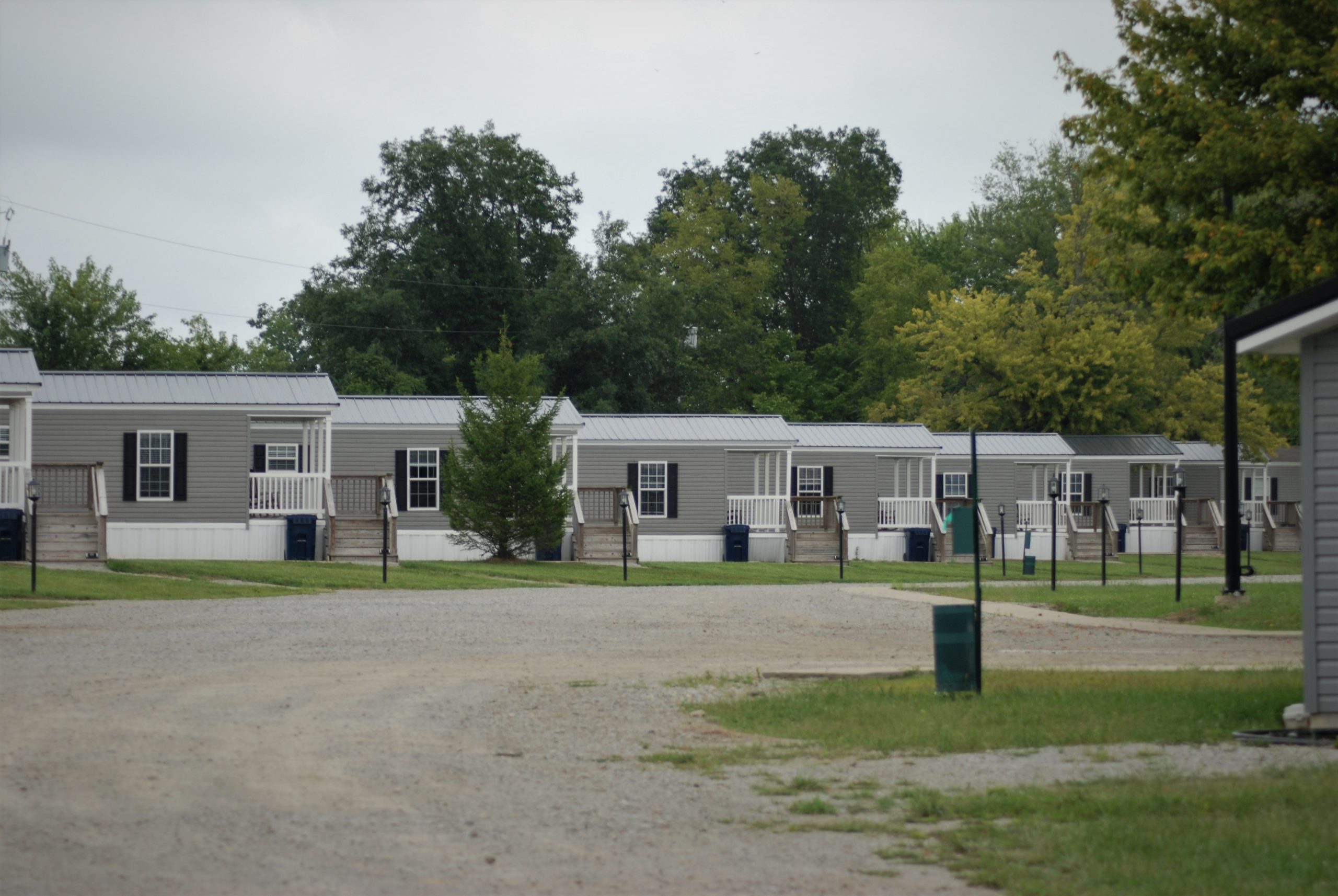 A row of well-kept manufactured homes with gray siding, white porches and skirting along a gravel lane in a manufactured housing community
