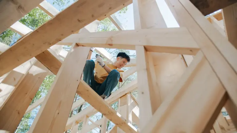 Carpenter in work gloves sitting on the wooden roof trusses of a building under construction