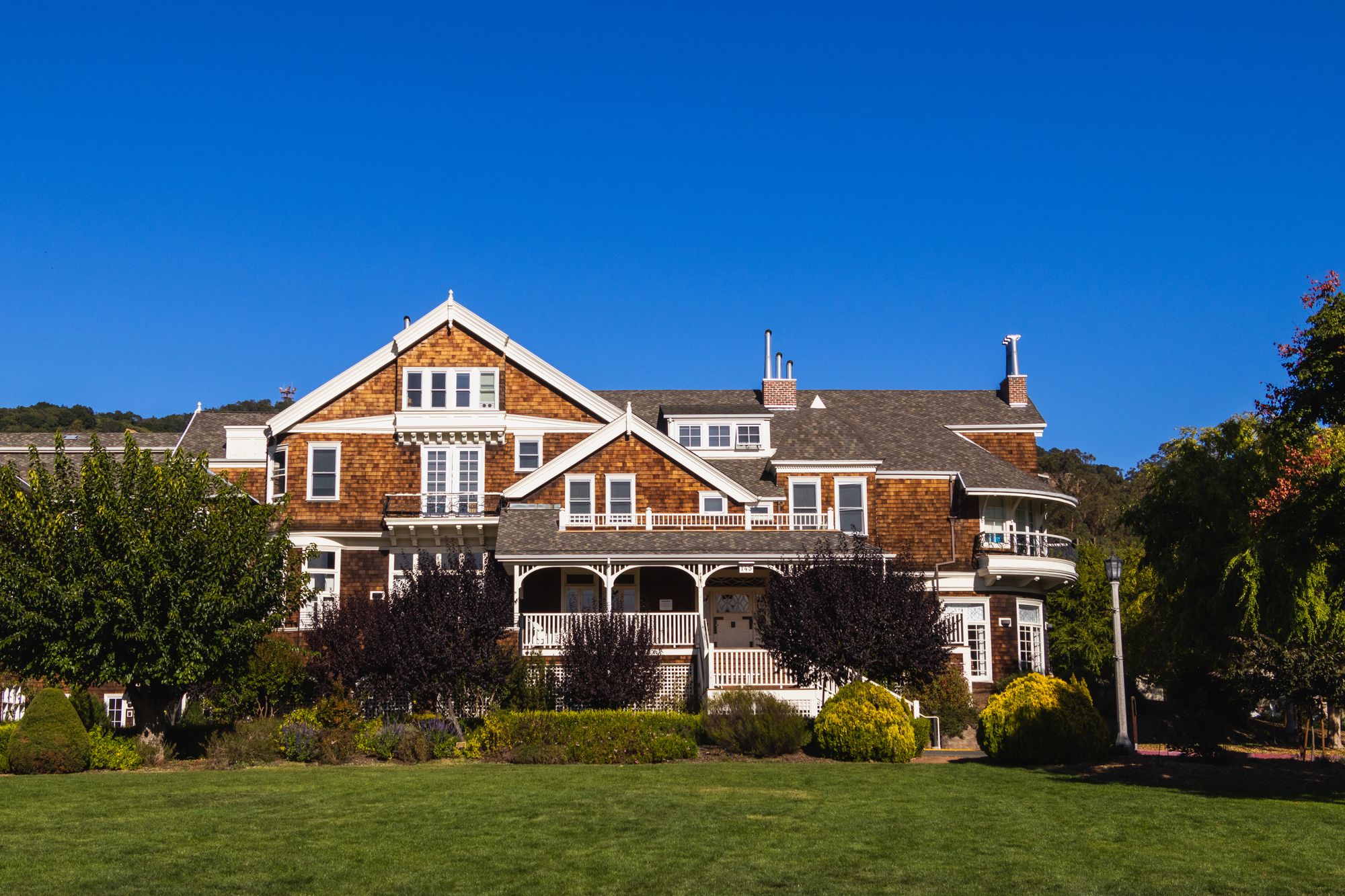 Large cedar shingle-style estate home with a covered porch, landscaped gardens and a wide manicured lawn under a clear blue sky