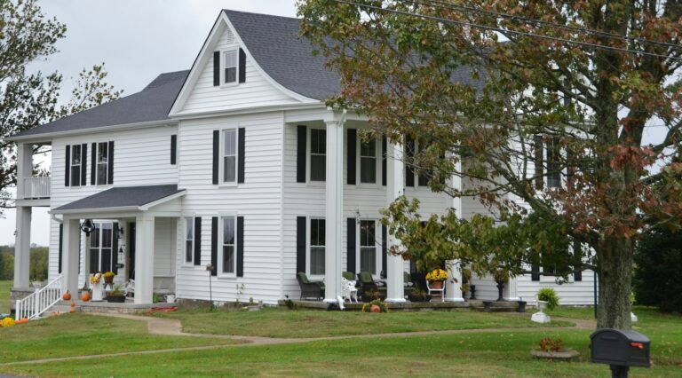 Large white two-story colonial farmhouse with black shutters, tall columned porches and a wide lawn in early autumn