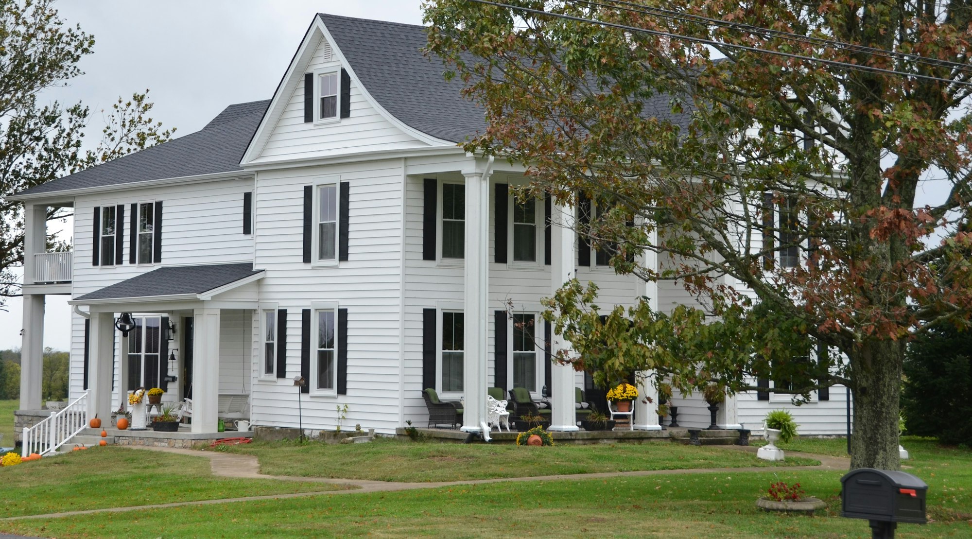 Large white two-story colonial farmhouse with black shutters, tall columned porches and a wide lawn in early autumn