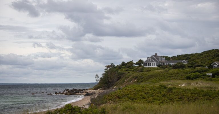 Gray-shingled coastal New England home on a bluff above a rocky beach under a cloudy sky
