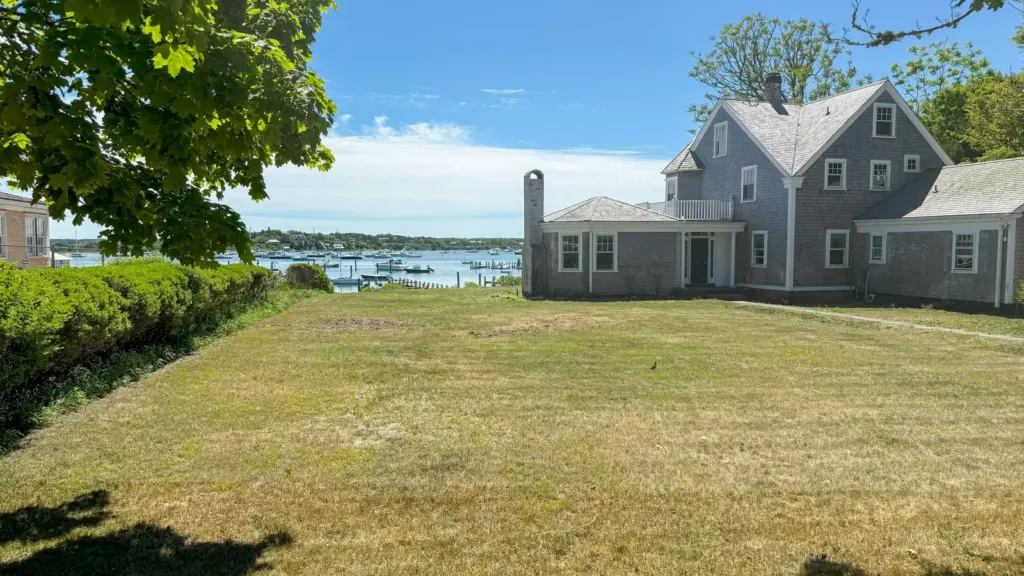 Gray cedar-shingled two-story house set back on a wide green lawn, with boats moored in Edgartown Harbor in the background on Martha's Vineyard, Massachusetts