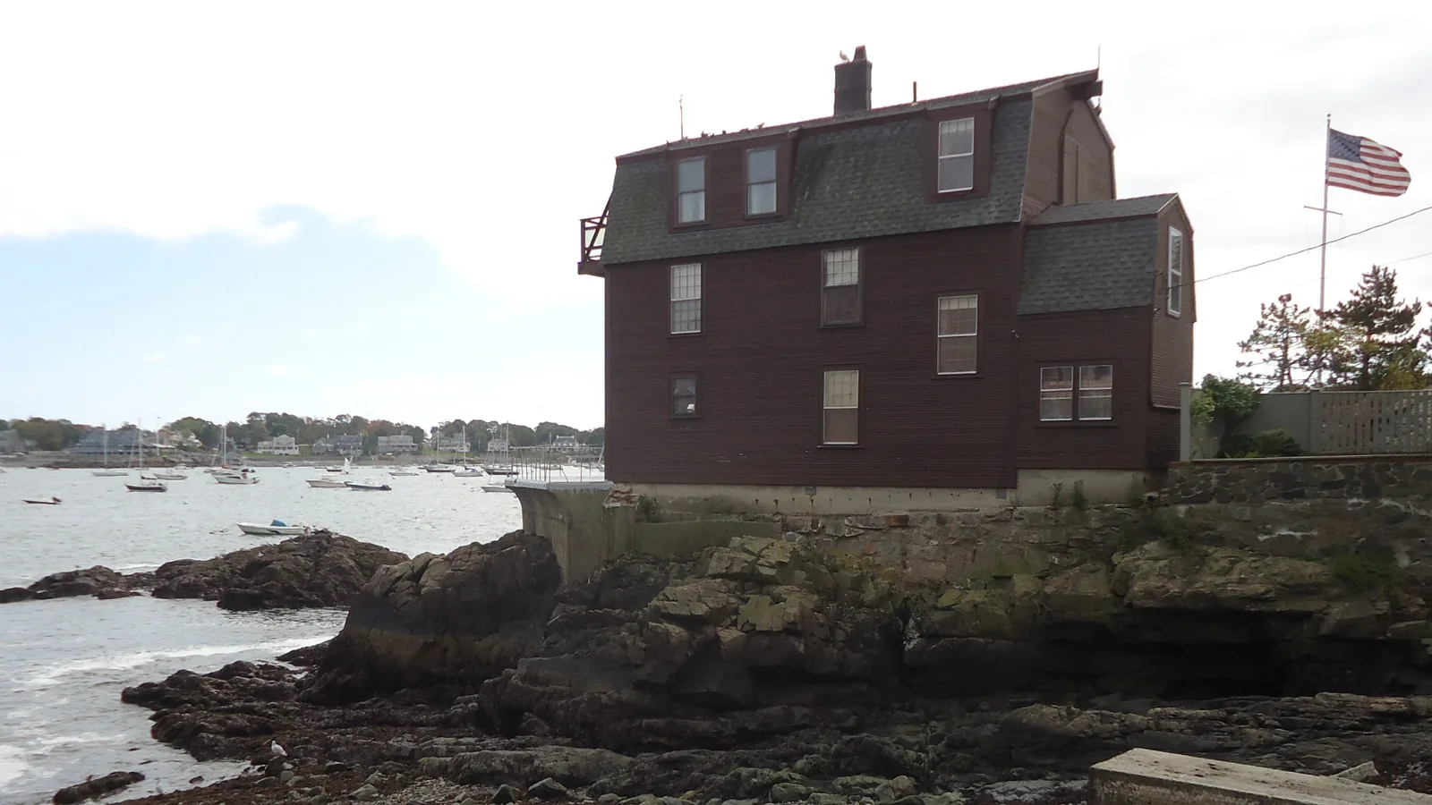 Older dark red-brown house with a steep roof, dormers and a chimney on a rocky ledge above a pebble beach at low tide, with moored boats in Marblehead harbor behind it