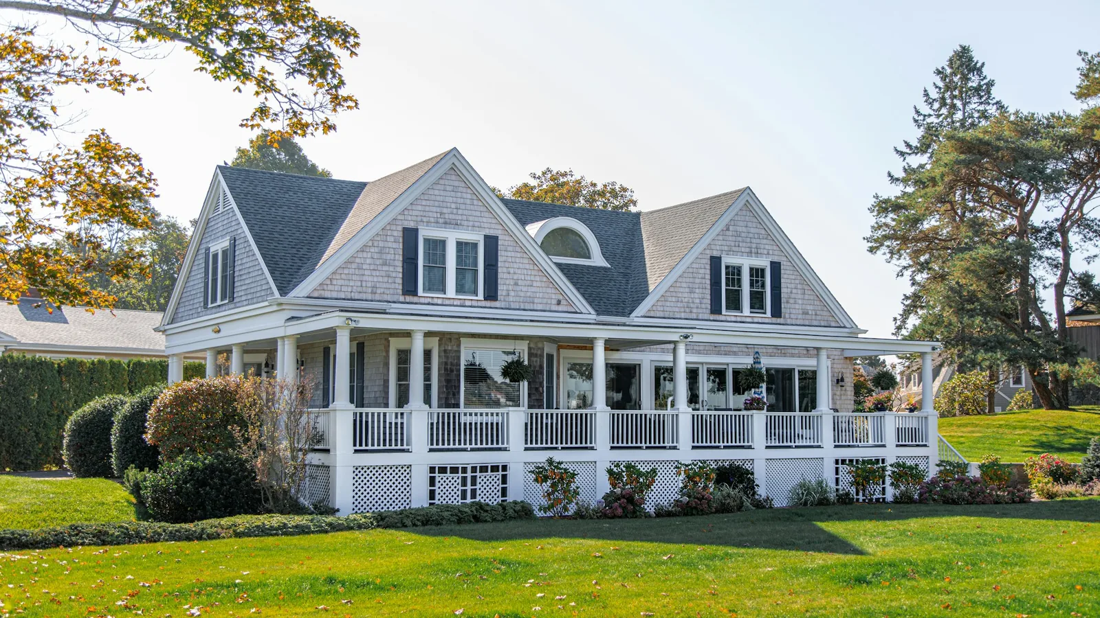 Gray cedar-shake Cape Cod-style home with a wraparound porch, white columns, and pine trees behind a green lawn in coastal New England